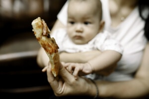 Mother is holding her baby on her lap and the other hand is holding a slice of pizza in front.
