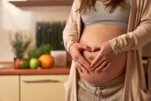 A pregnant woman is stroking her tummy in the kitchen. The mom-to-be is stroking her belly and holding a heart symbol. Fresh vegetables and fruits on the background of pregnancy.