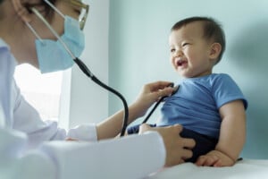A healthcare professional in a white coat and face mask uses a stethoscope to examine a smiling baby in a blue shirt for any signs of developmental delay. The baby is sitting on an examination table against a light blue wall.