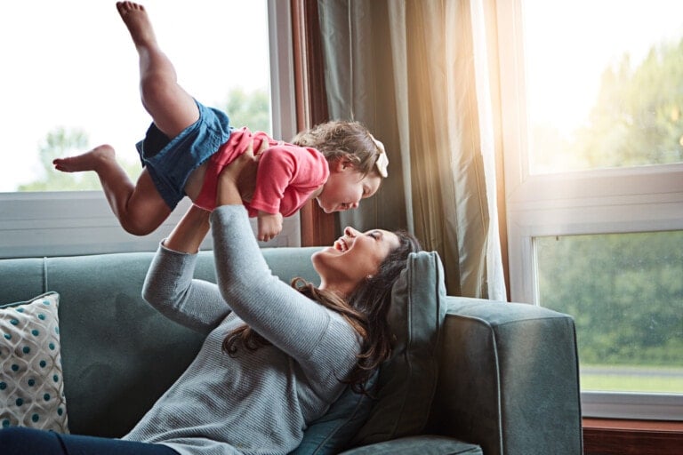 A woman is lying on a green couch holding a young child above her with both hands. The child, in a pink shirt and blue shorts, is smiling back at her. They are both beaming, showcasing the beautiful bonding between parents and children as sunlight streams through the window behind them.