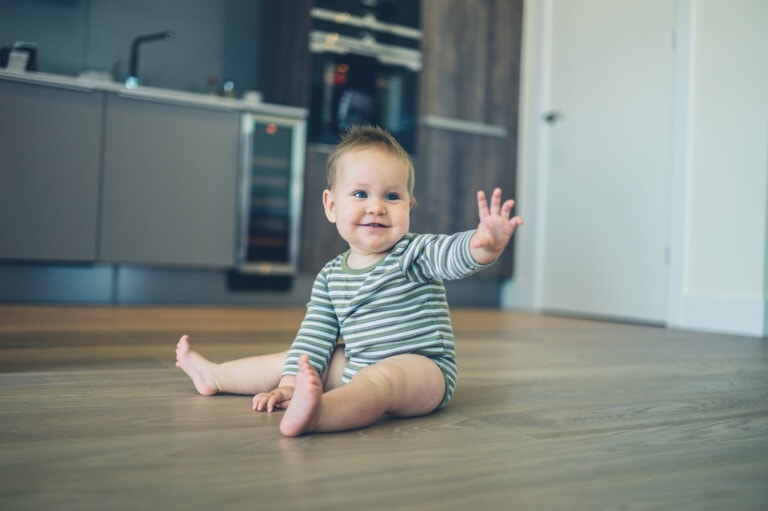 A cute little baby is sitting on the kitchen floor and is waving