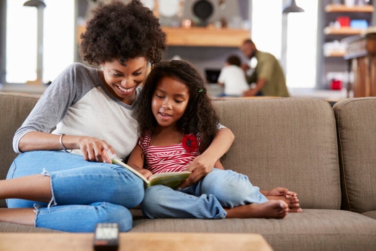 A woman and a young girl are sitting on a grey couch reading a book together. The woman is wearing a grey and white long-sleeved shirt and jeans, while the child sports a red and white striped sleeveless top with jeans. Two other people are in the background in the kitchen, perhaps showing what parents should not do for children by letting them enjoy their own moments of discovery.