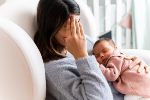 Mother sitting on couch in nursery feeling postpartum depression with baby in her arms. Many women suffer from postpartum blues after giving birth. In fact, it is estimated that 50-80% of mothers suffer from the 