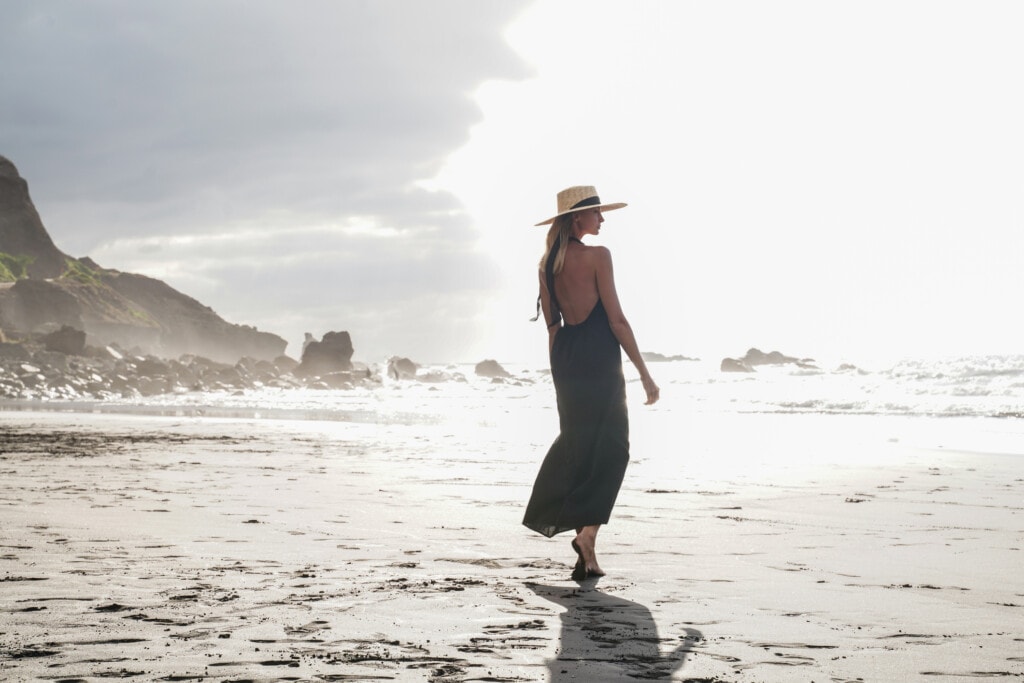 Beautiful romantic woman walking on the empty beach during sunset, wearing summer hat, looking at the ocean.