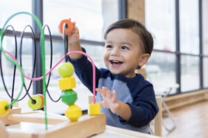 A toddler with short dark hair and wearing a blue long-sleeve shirt plays with a bead maze toy in a brightly lit room. Smiling and appearing engaged in the activity, the scene illustrates typical toddler behavior. Large windows are visible in the background.