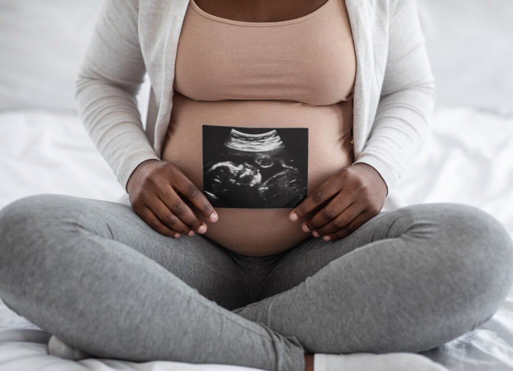 Unrecognizable black pregnant lady demonstrating her baby sonography photo while sitting on bed at home, showing firts photo of her child, enjoying happy maternity time, cropped image, closeup
