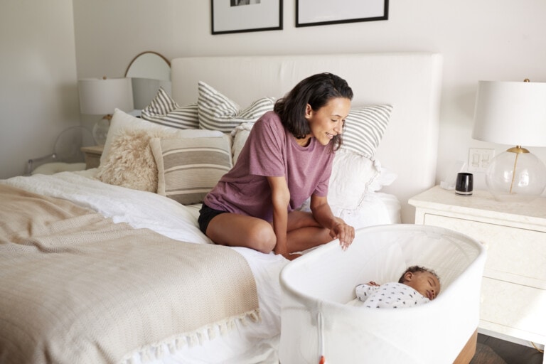 Young adult mother sitting on her bed looking down at her three month old baby sleeping in his cot