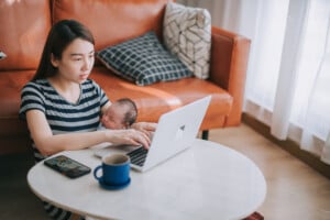Asian Chinese beautiful woman working from home using laptop carrying her baby boy in living room