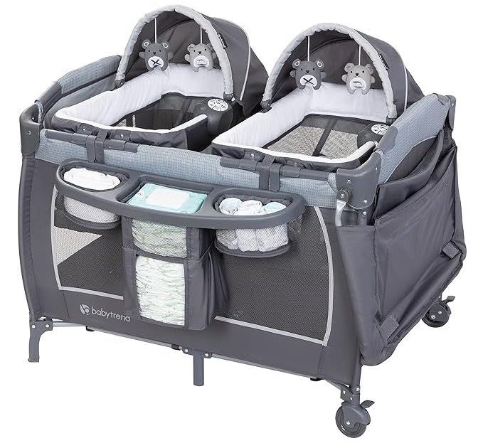A gray twin baby playpen with two attached bassinets, each featuring plush bear decorations. The playpen has a built-in diaper changing station, storage compartments for diapers and wipes, and is equipped with wheels for mobility. The brand "babytrend" is visible—one of the best baby products for twins. Baby Trend Lil' Snooze Deluxe III for Twins, Cozy Grey