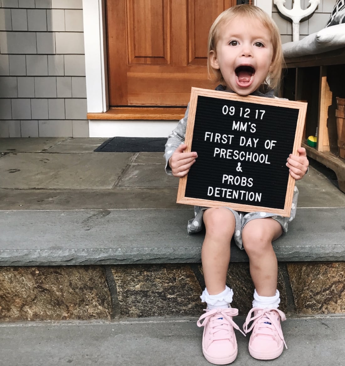 Little girl sitting on her first step holding up a letter board on her first day of preschool.