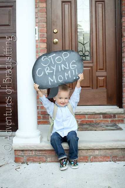 Little boy sitting on front porch step holding up a chalkboard sign that says "stop crying mom!"