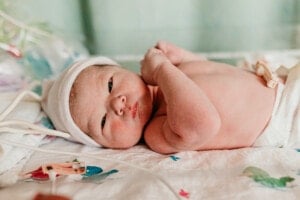 A newborn baby lying on a hospital bed with a white blanket, wearing a white hat with its eyes open and connected to medical tubes. The background shows medical equipment and a light green wall—a precious moment that highlights important photos to capture during baby's first 24 hours.