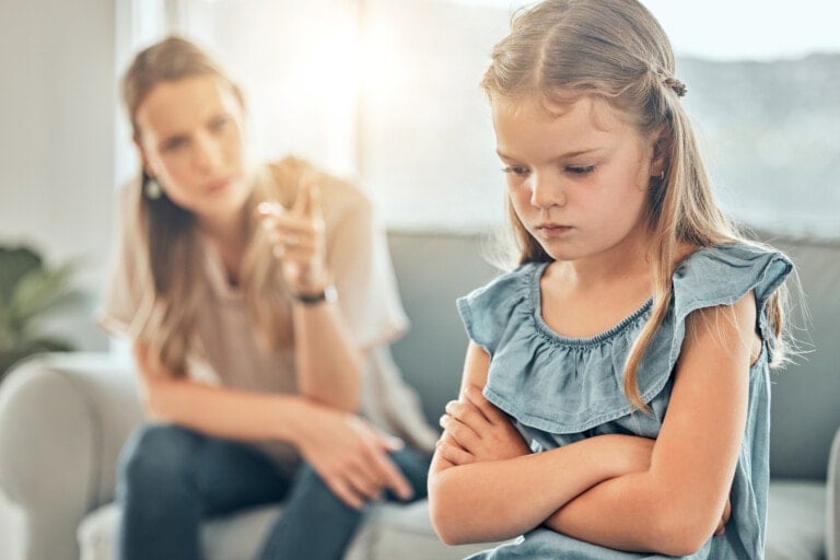 A young girl with blonde hair sits on a couch, looking upset and crossing her arms. An adult woman with blonde hair sits beside her, pointing and speaking to her, trying to discipline her stubborn child. The background includes a sunlit window.