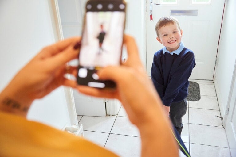 A young child in a school uniform smiles while standing in a hallway, perfect for first day of school picture ideas. An adult is taking a photo of the child with a smartphone. The child's image is visible on the phone screen, and there is a white door in the background.