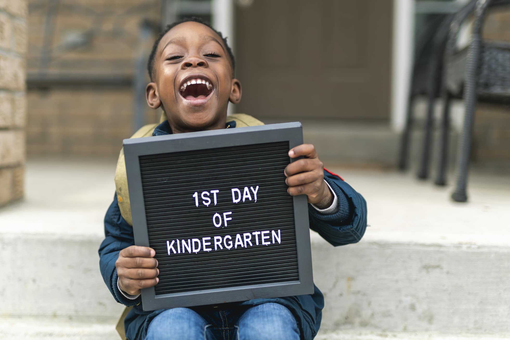 A cute boy of African America ethnicity sits on the patio stairs at his family home. He is holding a small black sign that reads 'first day of kindergarten'. The boy is laughing and very excited. He is wearing a backpack and winter clothes.