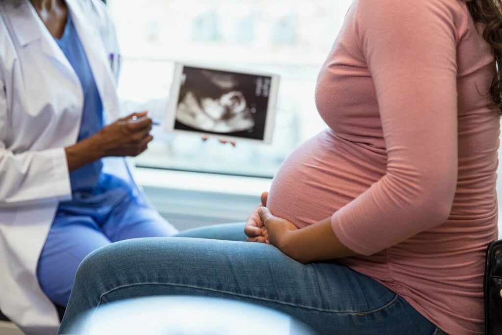 A photo with the focus on the unrecognizable pregnant woman in the foreground as the unrecognizable doctor shows her an ultrasound on a digital tablet in the background.
