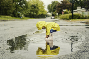 A Funny cute baby girl wearing yellow waterproof coat and boots standing in a big puddle and playing in the rain