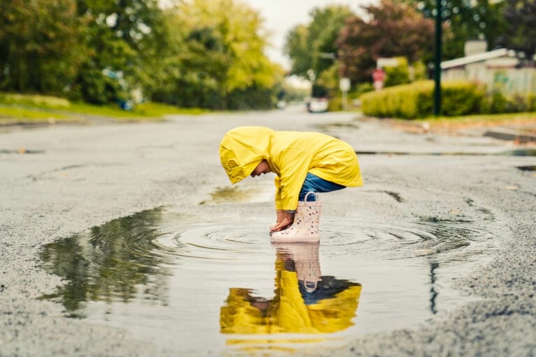 A child in a yellow raincoat and rain boots crouches down to touch a puddle on a wet street, reminiscent of old school parenting trends that encouraged outdoor play. The overcast sky and blurred greenery in the background indicate a recent rainfall.