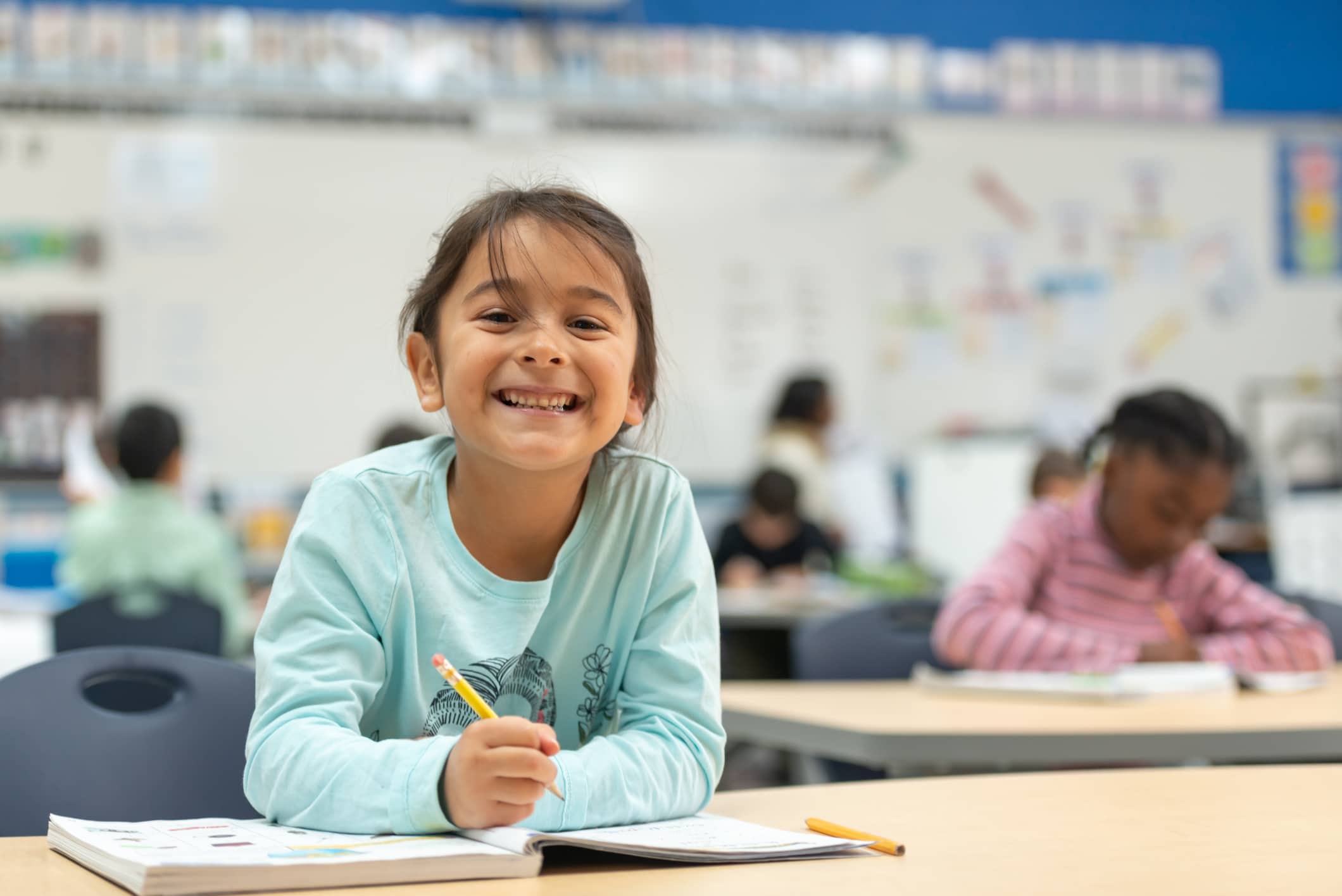 A cute little girl of spanish descent works hard on her in class assignment. She is working in her math workbook and smiling at the camera.