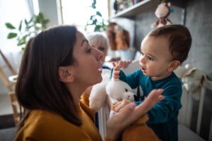 A woman and a baby interact indoors. The baby, holding a stuffed animal, reaches out to the woman, who is making a surprised expression. With plants and toys in the background, it's an ideal moment to get your child to start talking through playful engagement.