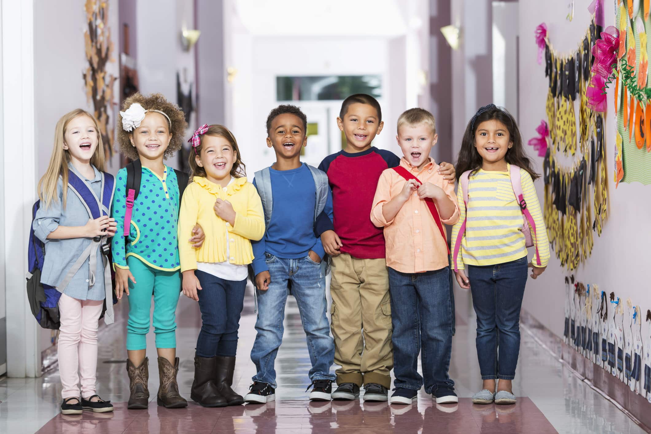 A multi-ethnic group of seven children standing in a row in a school hallway, laughing and smiling at the camera. The little boys and girls are kindergarten or preschool age, 4 to 6 years.