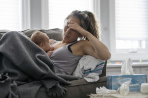 A middle aged mother resting on her couch holds her baby in her arm and holds her other hand over her head with a tired and stressed expression.