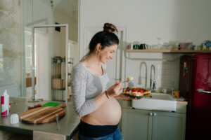 A pregnant woman with her hair in a bun is standing in a kitchen, eating for two from a plate of waffles topped with fruit and whipped cream. She is wearing a grey striped top and jeans. The kitchen has a red refrigerator, white sink, and various kitchen items.