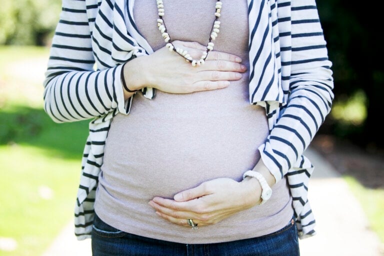A person standing outdoors, wearing a striped cardigan over a beige top and blue jeans. Their hands are placed on their pregnant belly, one above and one below, embraced in your second trimester. They are also wearing a beaded necklace and a watch on their left wrist.