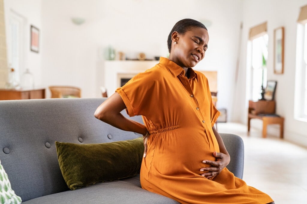 Mature pregnant woman having a backache while relaxing on couch at home. Unhappy black expecting woman suffering from lower back pain sitting on sofa with copy space. Tired middle aged african pregnant woman suffer from lower back pain on last month of pregnancy.