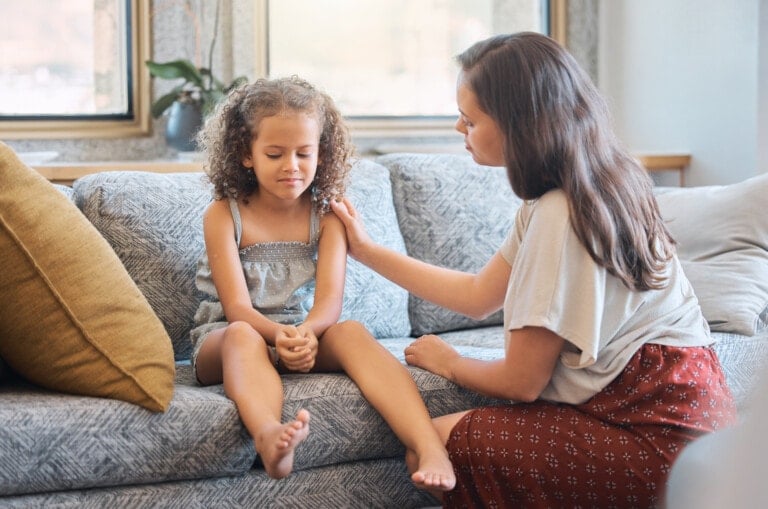 Sad little girl sitting on couch while mother tries to talk to her. Loving caring mother trying to communicate with upset daughter. Young hispanic mother asking little girl whats wrong while trying to comfort her and show support.
