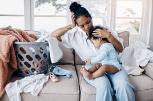 A woman sits on a couch, holding a toddler in her lap, their clinginess apparent. There is a laundry basket filled with clothes beside her and more clothes scattered on the couch. The woman rests her head on her hand, appearing tired. The room is well-lit with large windows in the background.