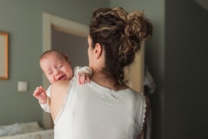 A person with curly hair in a bun holds a crying baby over their shoulder, perhaps wondering why babies cry when you sit. The person is wearing a white sleeveless top, standing in an indoor room with muted green walls and a doorway in the background.