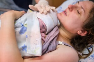 A woman lying in bed holds a newborn baby wrapped in a blanket on her chest. She appears to be in a hospital room, wearing a medical gown with her eyes closed, perhaps reflecting on the many things women say after childbirth. The baby is partially covered and wearing a cap.