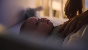 A baby lies in a crib, looking up, with an adults hands gently resting on their chest in a softly lit room.