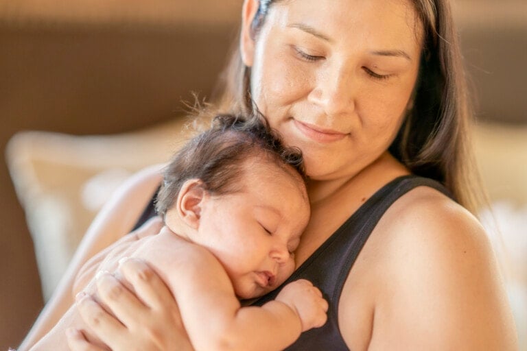 An adorable photo of a three (3) month old baby girl sleeping on her mother's chest. Her mother is looking down at her baby daughter with a soft smile on her face.