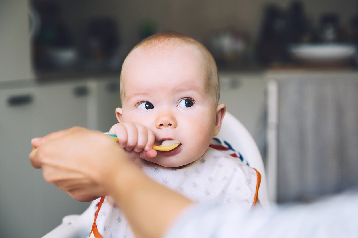 Feeding. Messy smiling baby eating with a spoon in high chair. Baby's first solid food. Mother feeding little child with spoon of puree. Daily routine. Finger food. Healthy child nutrition.