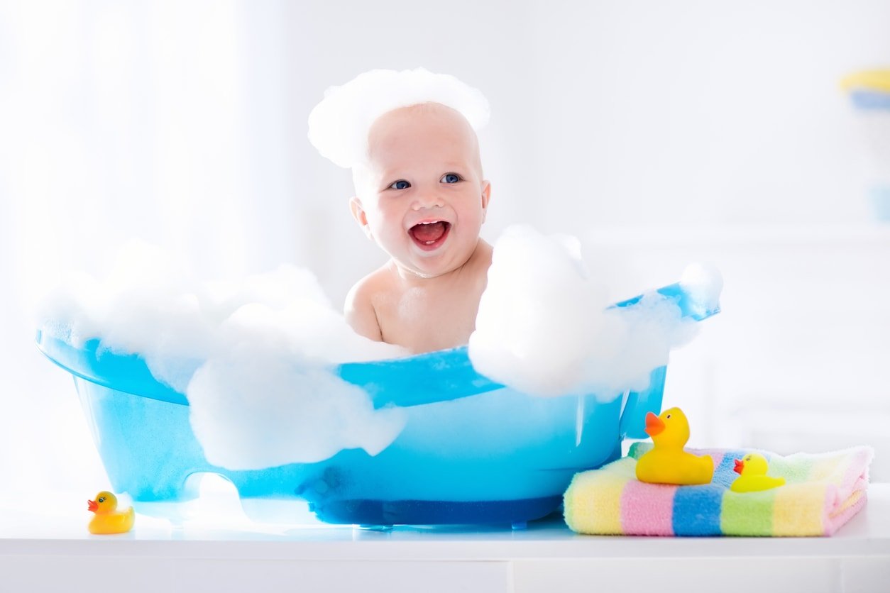 Happy laughing baby taking a bath playing with foam bubbles. Little child in a bathtub. Smiling kid in bathroom with colorful toy duck.