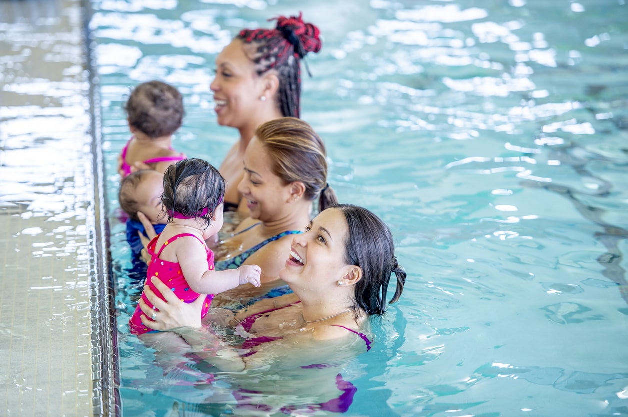 Three middle aged mothers hold their babies in a public pool during lessons.