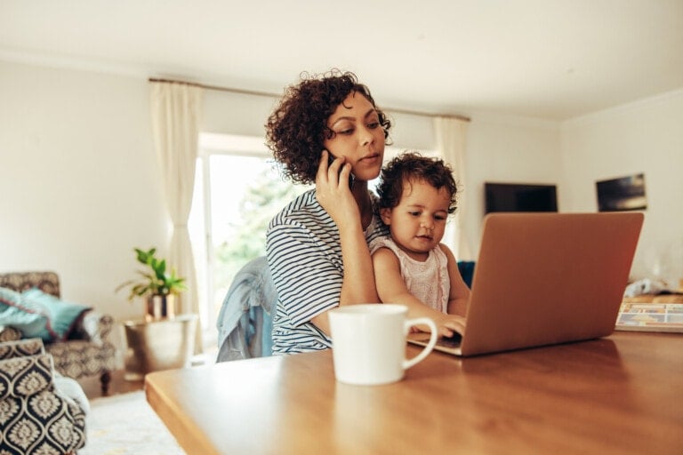 Woman freelancer using cellphone and laptop with her baby sitting on her lap at home. Working mother at home with baby.