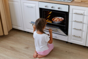 Portrait of little girl sitting near oven on floor in kitchen, open oven and looking at pie baked, kid posing backwards, wearing white t shirt and pink shorts.
