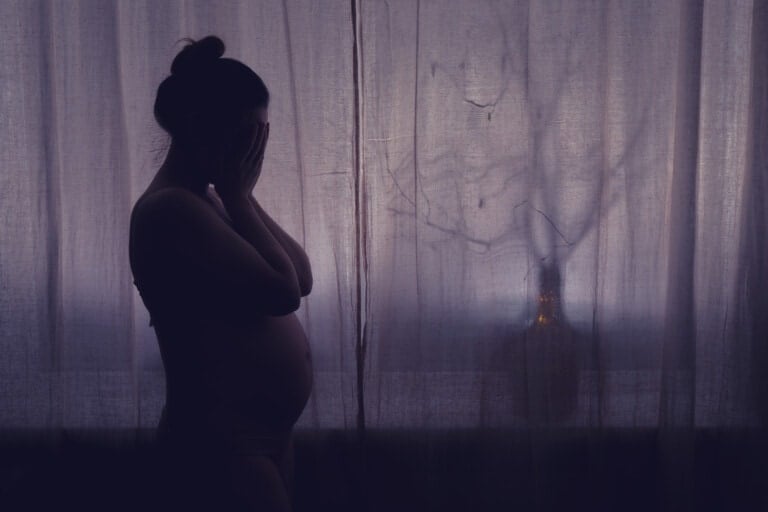 Young pregnant woman crying with her hands folded near her face. Pregnancy problems, silhouette of the expectant mother at the night window