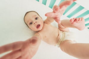 A baby lying on its back in a crib, wearing a diaper, and reaching out towards the camera with an expression of curiosity. The turquoise and white striped background adds a soothing touch. As you snap the photo, you might wonder, "Does my baby miss me?