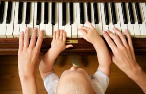 A baby and two adult hands are playing the piano. The baby's hands are placed on several keys, and the adult hands are positioned around the baby’s hands on the keyboard. This heartwarming scene of music for kids shows the baby standing in front of the piano, eagerly exploring its sounds.