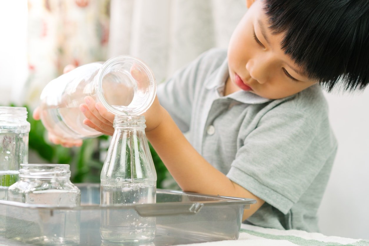 Cute Asian little Montessori boy pouring water into glass water bottle with concentration on wooden table at home