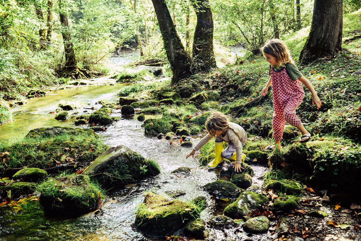 Two children having an adventure, climbing and stepping in a river exploring a beautiful natural area. Beautiful, idyllic and tranquil childhood moment. Sisters or friends, innocent and free.