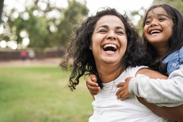 A woman and a young girl are outdoors in a grassy area. The woman is smiling broadly, and the girl, who is on the woman's back, is also laughing. Both have long, dark, curly hair. They appear to be enjoying the moment together as they explore the 5 love languages of children. Trees and blurred figures are in the background.