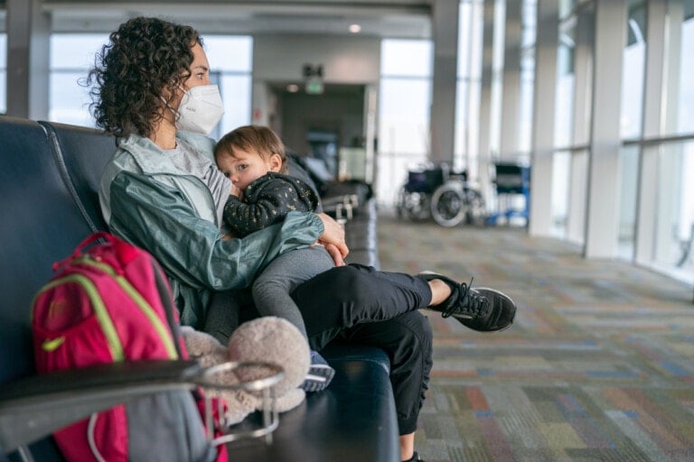 A young Eurasian mother wearing a protective face mask nurses her toddler daughter while waiting in the airport terminal to board their flight.