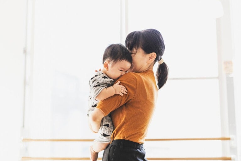 A woman is holding a baby in her arms, capturing one of those perfect baby-mom moments. She faces forward while the baby rests its head on her shoulder. With her hair tied back and dressed in an orange top, she stands against a bright indoor background.