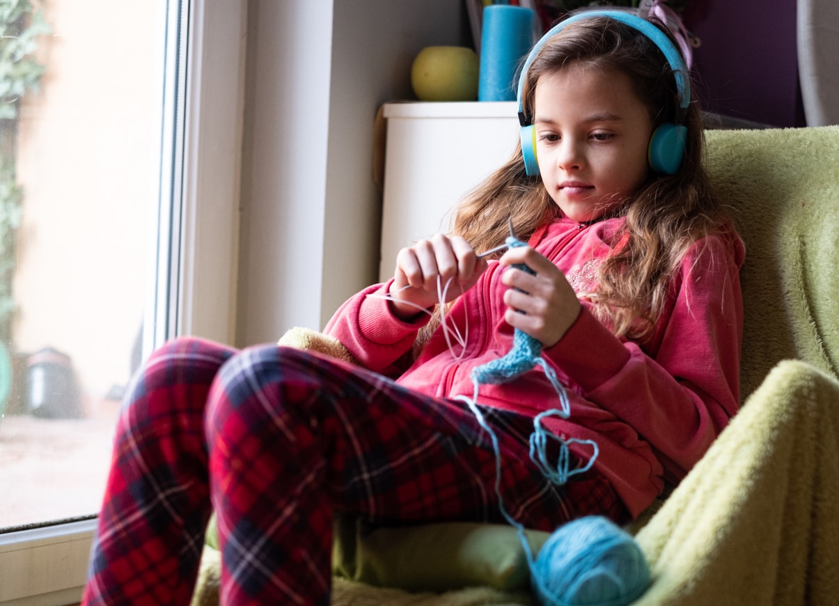 Small cute girl enjoying knitting at home while listening to music on her headphones.