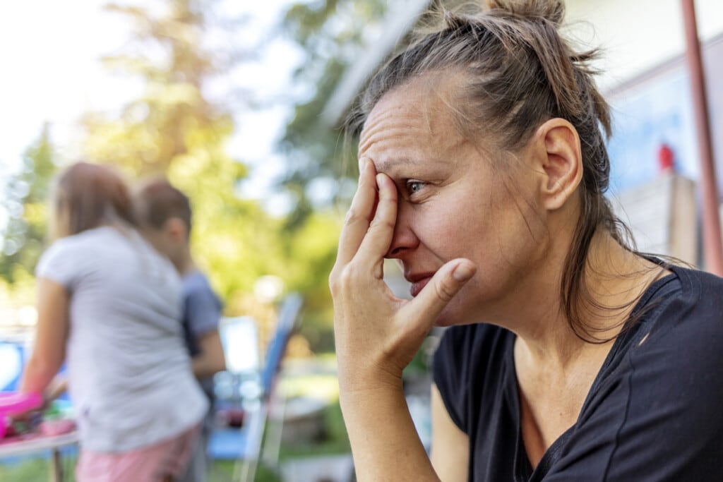 Stressed depressed young mother sits outside in a playground with her kids playing behind her. Single frustrated woman hold her head with hands sitting on chair with playful kids on a background.